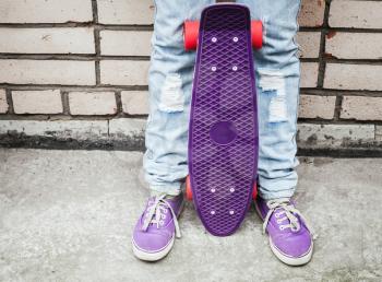 Teenager in jeans and gumshoes holds purple skateboard near by gray urban brick wall