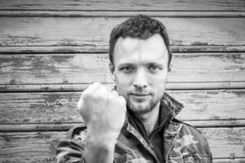 Young Caucasian man in camouflage showing his big fist. Monochrome outdoor portrait over rural wooden wall