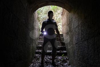 Young man with a flashlight enters stone tunnel and looks in the dark