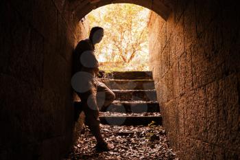 Young man stands in dark stone tunnel with glowing end, warm tonal correction filter