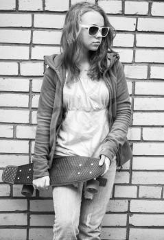 Teenage girl in jeans and sunglasses holds skateboard over urban brick wall background, monochrome photo