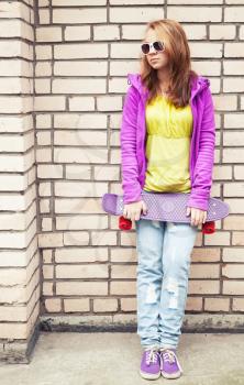 Blond teenage girl in sunglasses and colorful sporty clothes holds skateboard near by gray urban brick wall