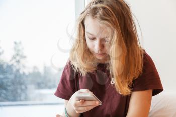 Blond teenage girl sitting near winter window with smartphone