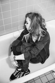 Stressed sad Caucasian blond teenage girl sitting in empty bath. Black and white vertical photo