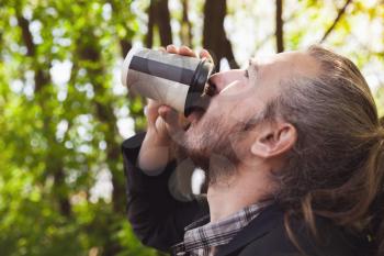 Bearded Asian man drinking coffee from paper cup in summer park, outdoor closeup profile portrait with selective focus