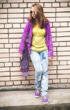 Blond teenage girl in sunglasses and colorful clothes with a skateboard near gray brick wall