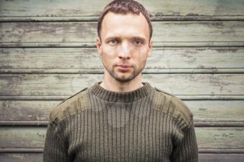 Young handsome Caucasian man in military green sweater. Outdoor closeup portrait over green rural wooden wall
