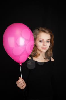 Studio portrait of smiling teenage Caucasian blond girl pointing with pink balloon over black background