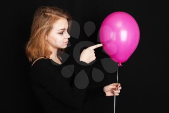Studio profile portrait of teenage Caucasian blond girl pointing with finger pink balloon over black background