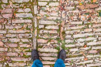 Male feet in leather shoes stand on old cobblestone pavement with grass ans fallen leaves, first person view