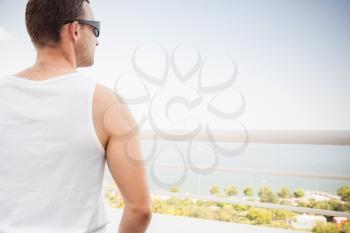 Young sporty Caucasian man in white shirt and sunglasses starring at the sea in bright summer day, rear view