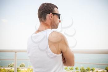 Young sporty Caucasian man in white shirt and sunglasses starring at the sea in bright summer day