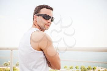 Young sporty Caucasian man in white shirt and black sunglasses. Outdoor summer portrait with sea on a background
