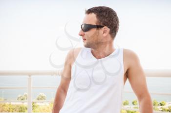 Young sporty Caucasian man in white shirt and sunglasses. Outdoor summer portrait with sea on a background