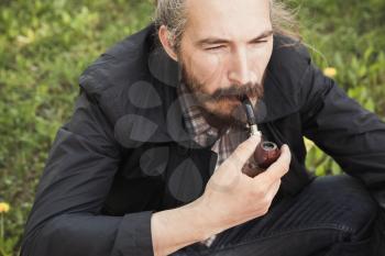 Asian man smoking a pipe on green grass in park, close-up portrait with selective focus
