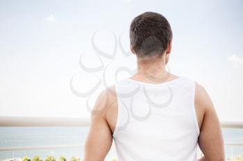 Young sporty Caucasian man in white shirt starring at the sea in summer day, rear view