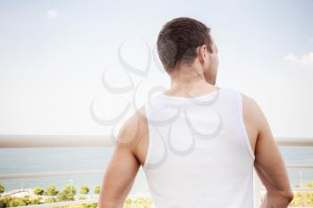 Young sporty Caucasian man in white shirt starring at the sea in bright summer day, rear view