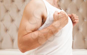 Young sporty Caucasian man torso in white shirt shows biceps in bedroom