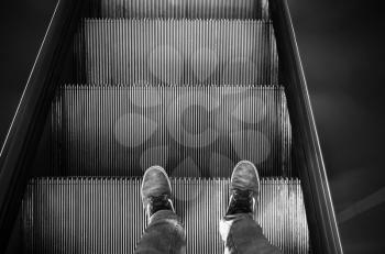 Male feet in canvas shoes stand on escalator stairs, black and white