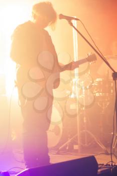 Rock and roll music background, blurred silhouette of electric guitar player on a stage with warm scenic illumination, soft selective focus