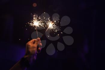 Two sparklers burn in one male hand over dark night background, soft selective focus