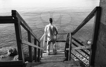 Young man stands on old wooden stairway going down to the sea coast, black and white photo