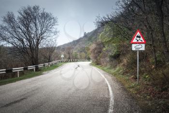 Slippery road, warning roadsign stands on mountain roadside, rainy rural landscape