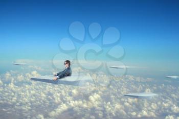 Businessman flying on big paper plane and wearing goggles isolated on sky background