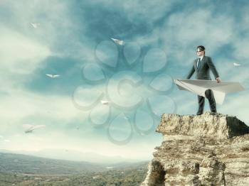 Businessman staying on mountain with big white paper plane and wearing goggles isolated on sky background