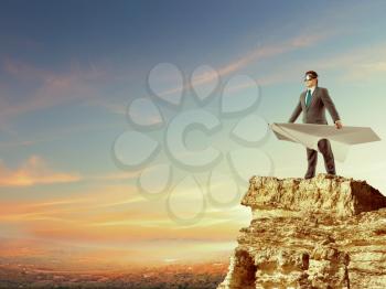 Businessman staying on the top of mountain with big white paper plane and wearing goggles isolated on sky background