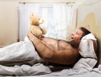 Adult man in bed looks at toy bear in home interior