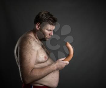 Side view of fat man looking at the sausage in his hand over grey.  Potency problem