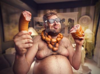 Happy man in glasses with sausages round his neck holds big sausages in both hand sitting in the room