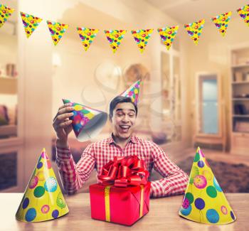 Young smiling man playing with Birthday hats and a present at the table