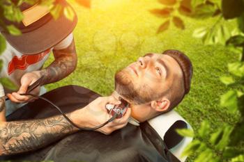 Young man having his beard shaven, barber working with trimmer over nature background