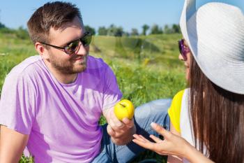Smiling couple eating apples while having picnic on the meadow