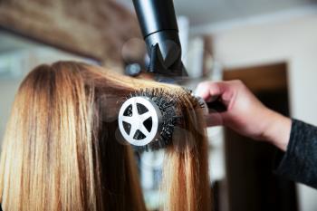 Professional woman hairdresser making hairstyle using hair dryer for young female in beauty salon.