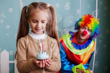 Smiling little girl holds cake in hand and makes a wish. Funny clown on the background.