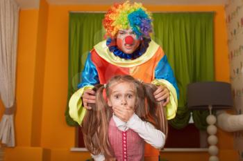 Little girl was frightened of the clown who has crept behind. Kindergarten with colorful couch on the background.