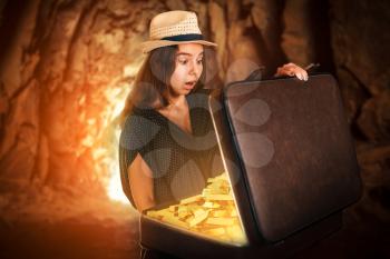 Young woman stands in a cave and holding a half-open suitcase full of gold bars. Vintage style. Adventure concept.