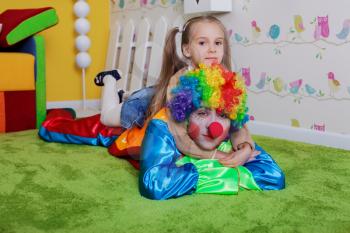 Funny girl sits on a back of the clown with red nose. Colorful sofa, nesting box and fence on the background. 