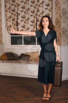 Young woman standing with suitcase and show hitchhiking sign. Vintage travel waiting concept. Sofa and window with curtains on the background.
