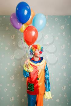 Dull clown with a bunch of colourful air balloons standing in the room.
