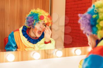Cheerful clown does a make-up in front of the mirror. Makeup room at the background