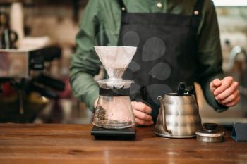 Metal coffee pot and glass on a bar counter against waiter in black apron. Blury coffee house on background.