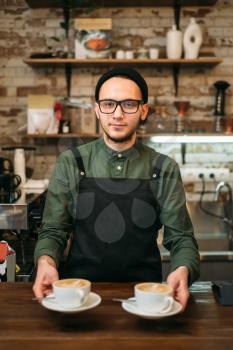 Waiter in black apron holds two cups of coffee in his hands.