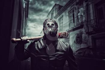 Angry psycho man in hockey mask and black leather coat with bloody baseball bat with a chain wrapped around. Maniac waiting for his victim on night city street.
