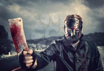 Butcher in hockey mask holding bloody meat cleaver in hand. Night road on background.