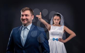Sly bride brought a finger gun at the back of her future groom, studio photo shoot, black background. 