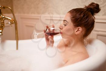 Beautiful nude woman drinking red wine in the bathtub with foam.